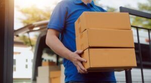 Delivery worker in a blue shirt carrying stacked cardboard boxes beside a van, illustrating commercial transport cases handled by H&P Accident & Injury Lawyers